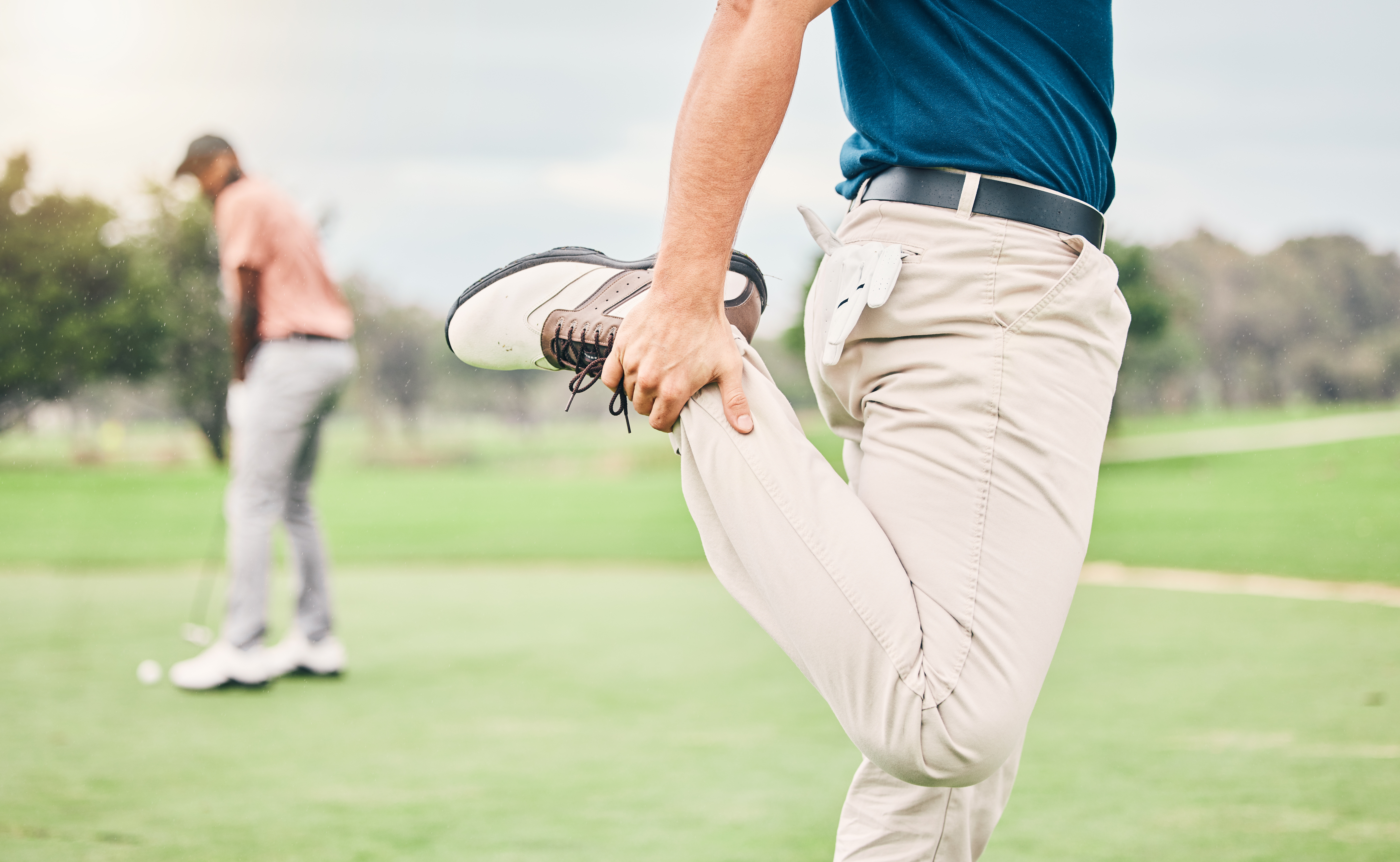Échauffement golf : golfeur effectuant un étirement du quadriceps sur le parcours, avec un autre joueur en arrière-plan