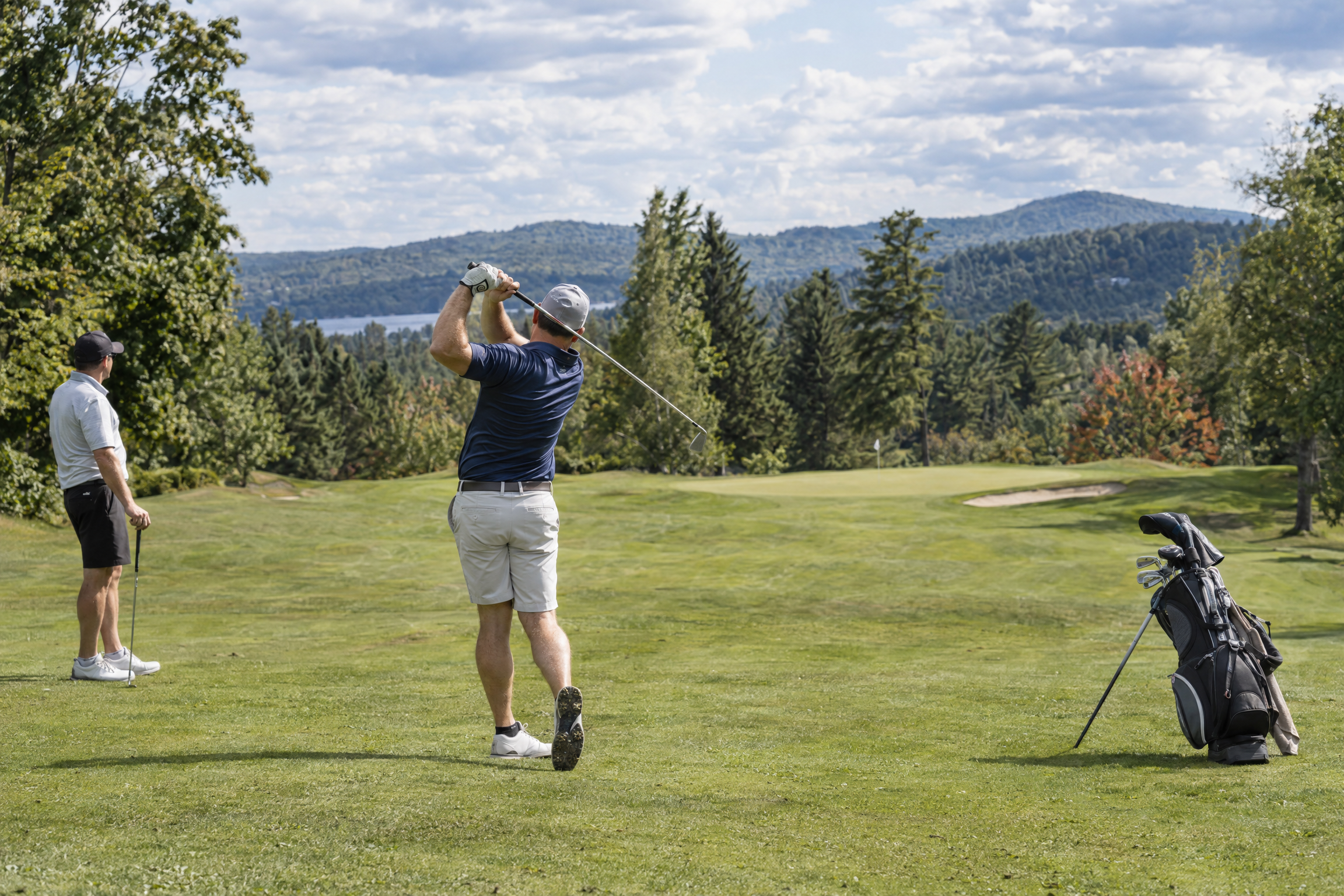 Golfeur en plein élan sur un fairway avec vue sur les montagnes, sous le regard de son partenaire qui suit sa progression et son handicap au golf.